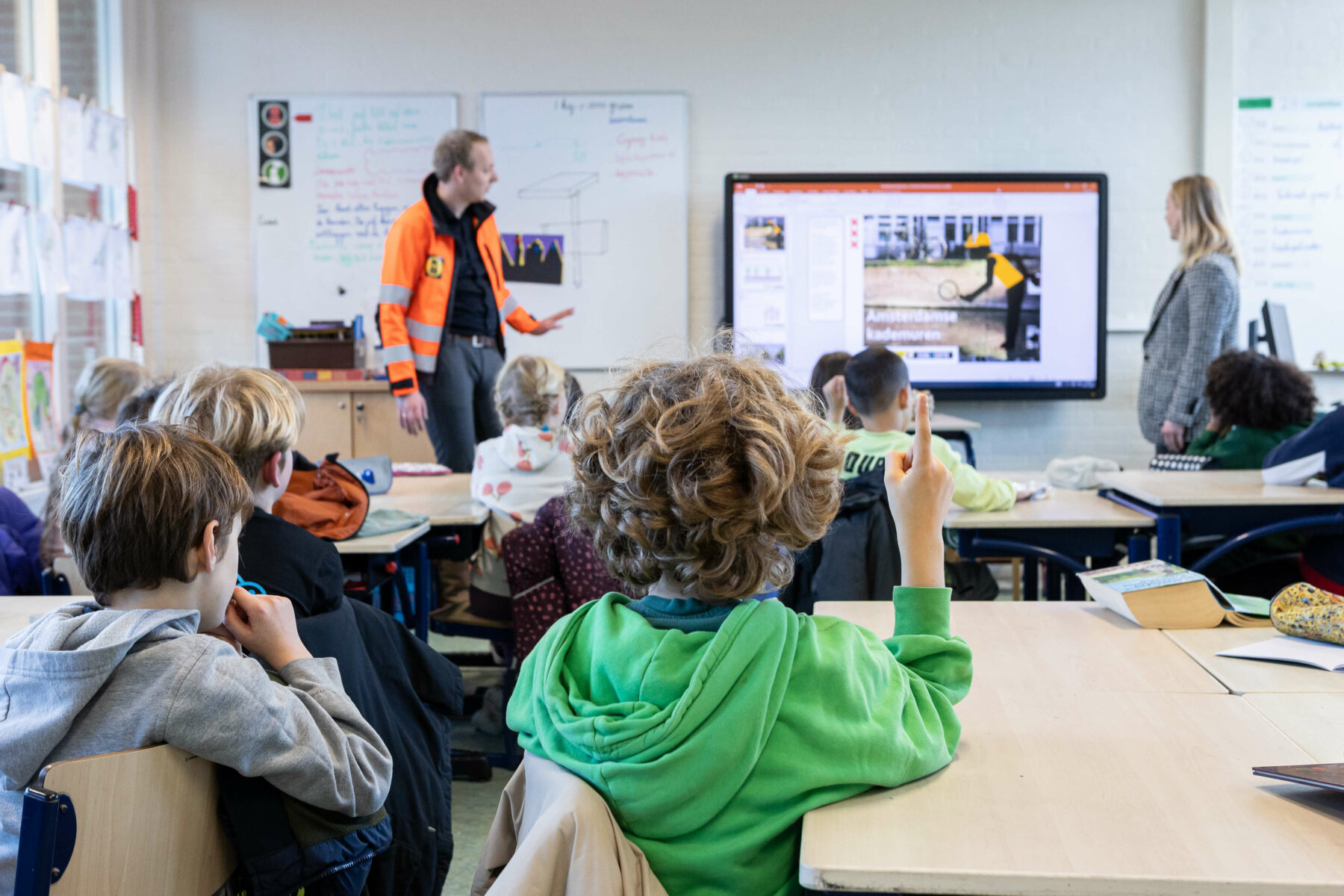 Jaap en Elise geven een presentatie in een klaslokaal vol leerlingen. Een leerling steekt zijn vinger op.