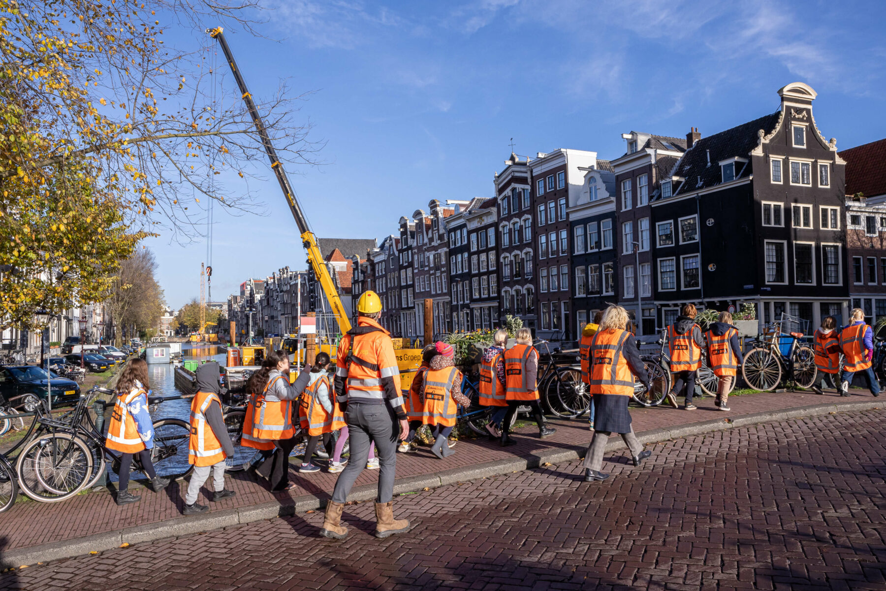 Een groep leerlingen in oranje veiligheidshesjes loopt over een brug en kijkt naar de werkzaamheden aan de gracht.